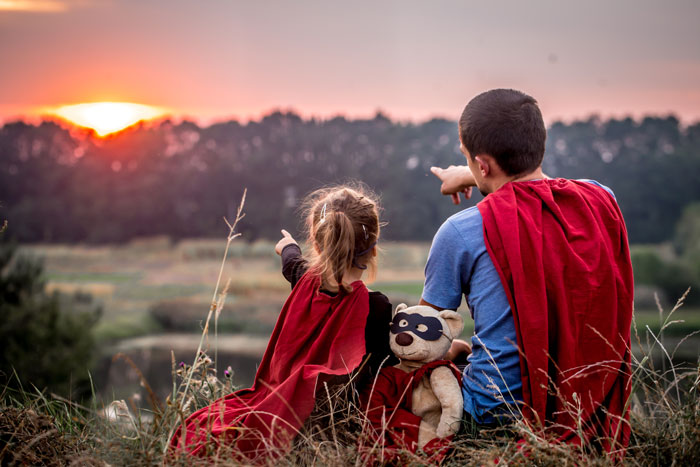 a father and son wearing capes sitting on the grass and watching the sunset